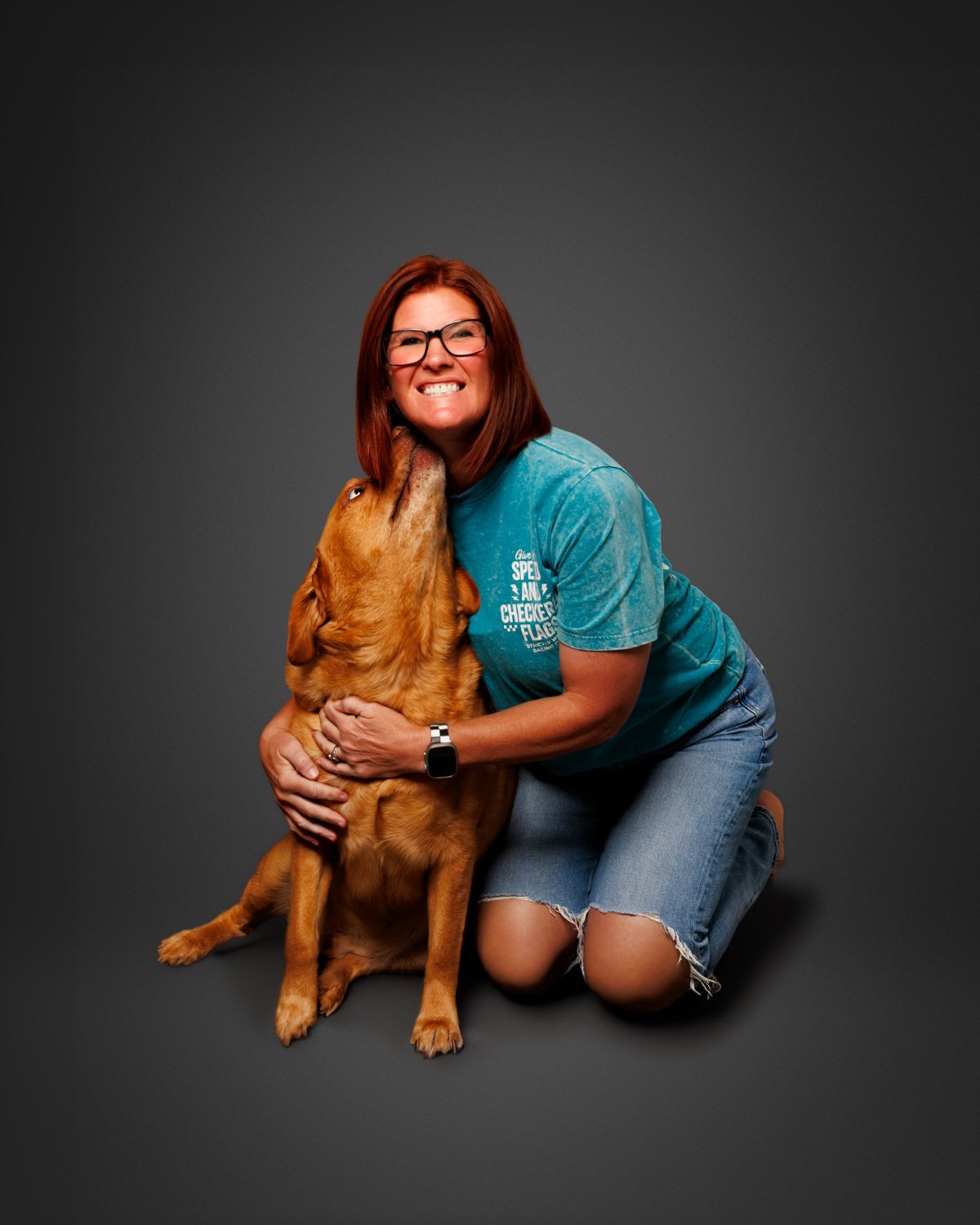 Amy Scotti kneeling on a gray studio backdrop while a golden lab licks her chin