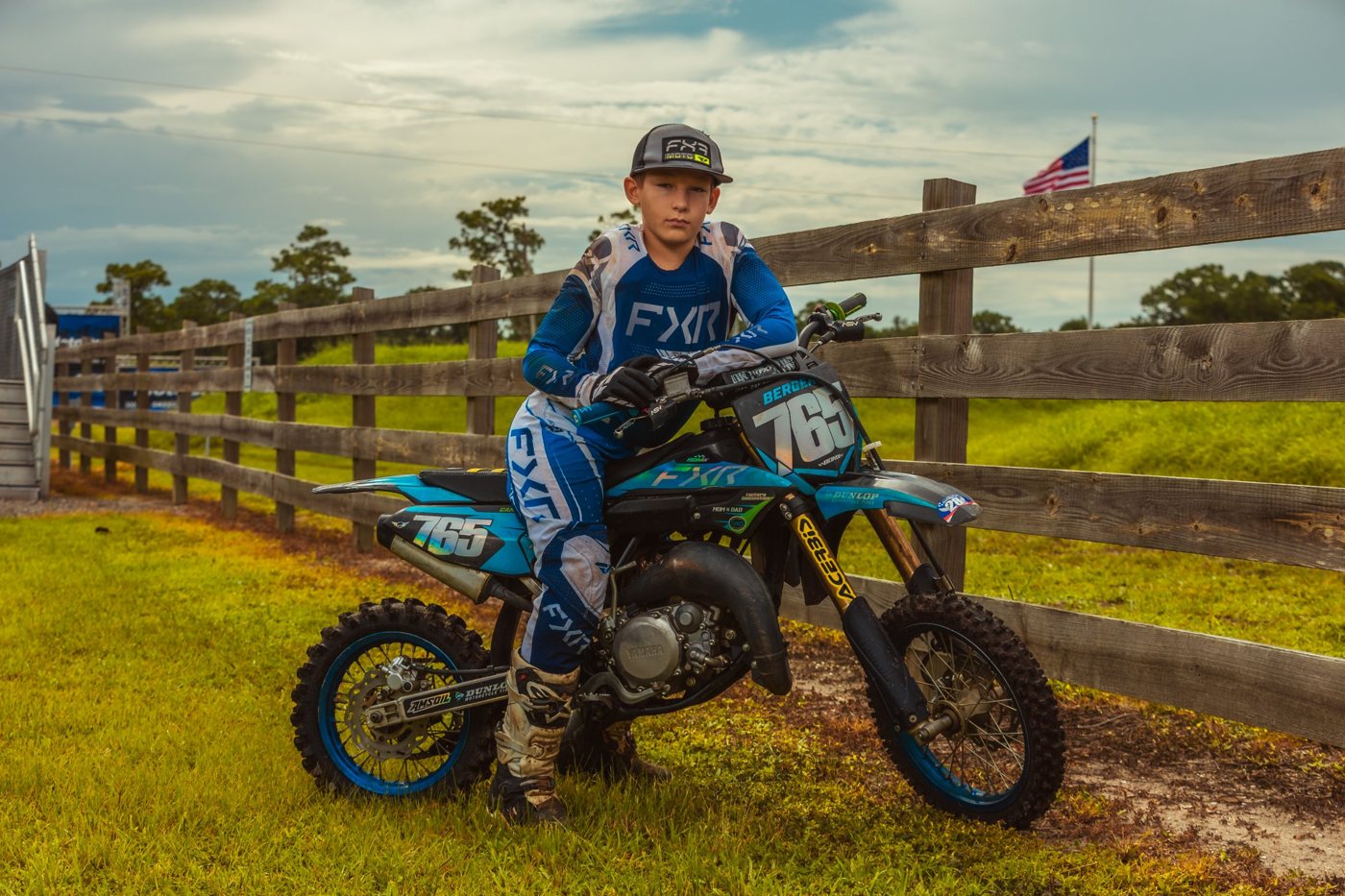 Young motocross rider in blue gear leaning on his bike beside a wooden fence at the track
